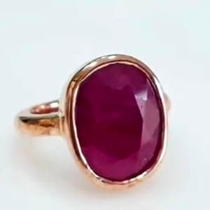 Könnte beinhalten: Ein roségoldener Ring mit einem großen, oval geschliffenen Rubin. Der Rubin hat eine tiefe, satte rote Farbe und ist in einer Zargenfassung gefasst. Der Ring hat ein schlichtes, elegantes Design.