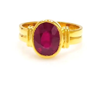 Puede incluir: Un anillo de oro con una gran gema de rubí de corte ovalado. El rubí es de color rojo intenso y está engastado en un bisel de oro. El anillo tiene un diseño simple pero elegante, con una doble banda y detalles dorados decorativos.