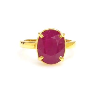 Puede incluir: Un anillo de oro con una gran gema de rubí de corte ovalado. El rubí es de un color rojo intenso y vibrante, y la banda de oro tiene un acabado liso y pulido. El anillo está sobre un fondo blanco.