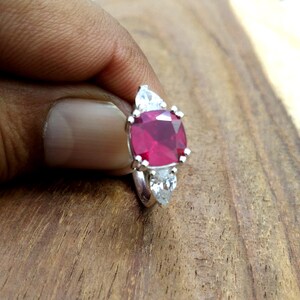 Puede incluir: Un anillo de plata con una gran gema de rubí de corte cuadrado, flanqueada por dos gemas blancas en forma de pera. El rubí es de color rojo intenso y el anillo está sobre una superficie de madera.