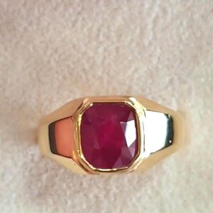 Op de afbeelding: Een gouden ring met een grote, rechthoekig geslepen robijn. De robijn heeft een dieprode kleur en is gezet in een gepolijste gouden band. De ring heeft een klassiek, elegant ontwerp.