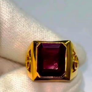 Puede incluir: Un anillo dorado con una gran gema rectangular de color rojo oscuro. El anillo tiene una banda gruesa y elementos decorativos en los laterales. La gema es el punto focal, engastada en un bisel dorado.