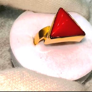 May include: A gold ring with a triangular, bright red gemstone. The ring's band and setting are polished gold, contrasting with the vibrant red stone. The ring is set on a white surface.