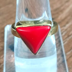 May include: A gold-toned ring featuring a vibrant red triangular stone. The ring's band and setting have a polished finish. The red stone is the focal point, creating a bold and eye-catching design. The ring is displayed on a clear, cylindrical stand.
