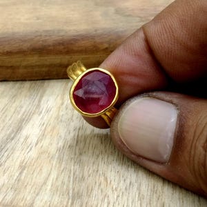 Puede incluir: Un anillo de oro con una gran gema de rubí ovalada y facetada. El rubí es de color rojo intenso y está engastado en un bisel de oro. El anillo tiene un diseño sencillo y elegante.