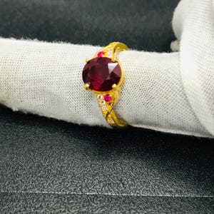 以下が含まれることがあります： 大きな楕円形の深紅色の宝石を中央に配したゴールドリング。リングは、詳細で装飾的なデザインにセットされた、より小さな赤と透明の宝石で飾られています。リングは白い布の上に展示されています。