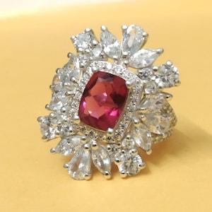 Peut inclure: Une bague argentée ornée d'une grande pierre précieuse rectangulaire rose foncé au centre. La bague est entourée d'un halo de pierres plus petites, claires, rondes et en forme de poire, créant un design étincelant en forme de fleur. La bague est posée sur un fond jaune.