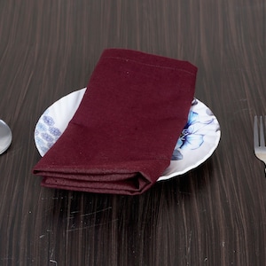 Puede incluir: Una servilleta burdeos doblada descansa sobre un plato blanco con un diseño floral azul. Una cuchara y un tenedor plateados están a cada lado del plato, sobre una mesa de madera oscura. La escena sugiere un entorno de comedor formal.