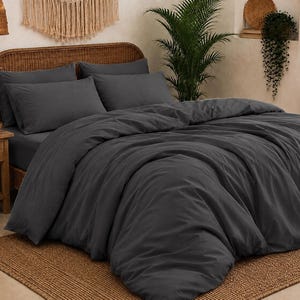 May include: A dark charcoal gray bedding set consisting of a duvet cover and two matching rectangular pillowcases. The fabric has a smooth, matte finish and is draped over a bed frame, creating soft folds and a layered appearance across the surface.