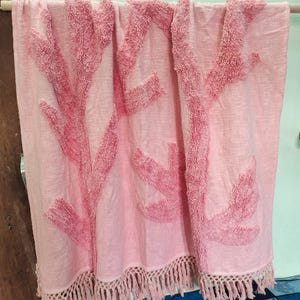 May include: A light pink throw blanket with coral-like, textured designs in a darker shade of pink. The blanket has a fringe trim along the bottom edge. The throw is hanging on a wooden rod.