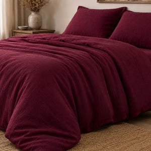 Peut inclure: Une parure de lit bordeaux profond comprenant une housse de couette texturée et deux taies d'oreiller assorties. Le tissu présente un aspect tissé et froissé visible, offrant un rendu doux et superposé sur toute la surface du lit en rouge foncé.