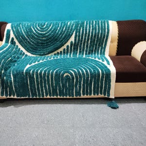 Op de afbeelding: Een turquoise en wit patroon deken over een bruine en beige bank. De deken heeft een herhalend boogontwerp met verticale lijnen. De bank heeft bruine kussens en beige armleuningen. Kwasten hangen aan de hoeken.