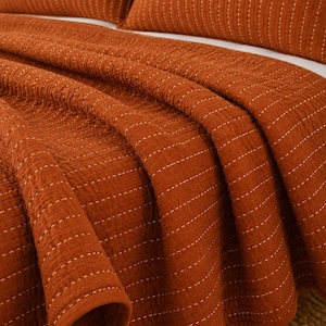 May include: A burnt orange quilt featuring a textured, crinkled fabric surface with vertical rows of white dashed stitching. The quilt is draped over a bed, showing a matching pillow sham in the background with the same stitched pattern.