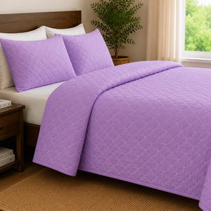 May include: A lavender quilt set featuring a diamond-stitched pattern across the bedspread and two matching pillow shams. The bedding is a solid light purple colour with a textured quilted finish, providing a uniform look for the bed setup.