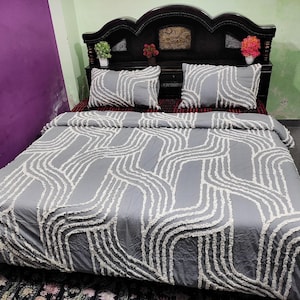 May include: A grey bedspread and two matching pillowcases with a white wavy pattern. The bedding is on a dark wooden bed frame with a decorative headboard. The room has a purple wall.