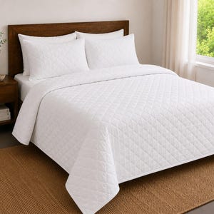 Puede incluir: Una colcha blanca y dos fundas de almohada a juego con un patrón acolchado en forma de diamante. El juego de cama se muestra sobre una cama con un cabecero de madera oscura, creando un aspecto limpio y uniforme para el área de descanso.
