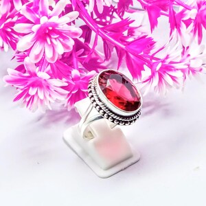 May include: A silver ring with a large, oval-shaped, red gemstone set in a silver bezel. The ring has a decorative, beaded border around the gemstone.