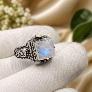 May include: A silver ring featuring a square-cut moonstone with a blue iridescent sheen. The band is crafted with a dark, oxidized finish and displays embossed star motifs on the sides, with a hinged box design holding the central gemstone.