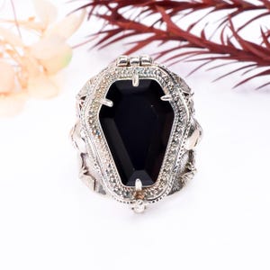 May include: A silver ring with a black coffin-shaped stone in the center. The ring is detailed with skulls and other gothic designs.