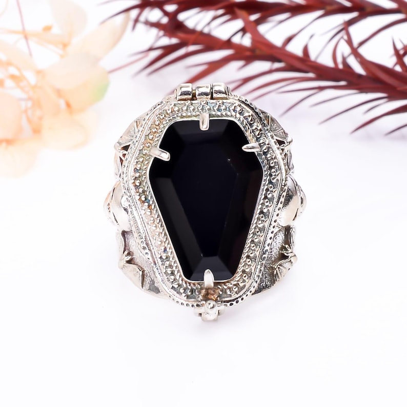 May include: A silver ring with a black coffin-shaped stone in the center. The ring is detailed with skulls and other gothic designs.
