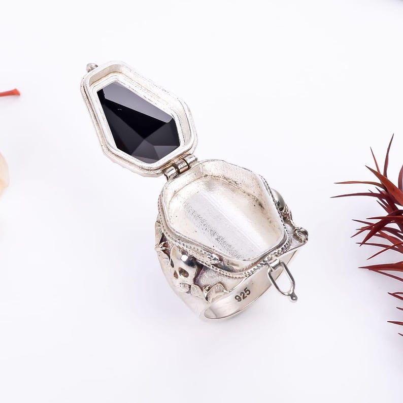 May include: A silver ring with a hinged compartment, decorated with skulls and a black gemstone. The ring is marked "925" on the inside.