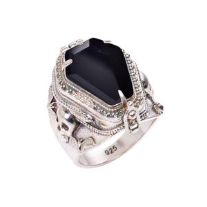 May include: A silver ring with a black gemstone set in a detailed, ornate design. The ring features a skull motif and the inscription "925" on the band.