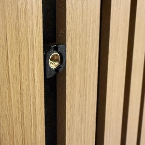 Op de afbeelding: Close-up van houten panelen met verticale, lichtgekleurde houten latten. Een zwarte beugel met een goudkleurige schroefdraadinzet is zichtbaar tussen twee latten. De houtnerf is prominent aanwezig.