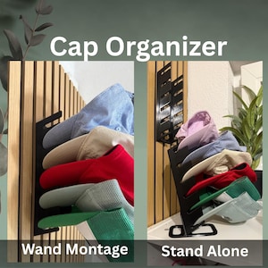 Puede incluir: Organizador de gorras de metal negro con múltiples gorras de béisbol en varios colores, incluyendo azul, beige, rojo y verde. El organizador se muestra en dos configuraciones: montado en la pared y de pie. El texto en la imagen dice "Cap Organizer", "Wand Montage" y "Stand Alone".