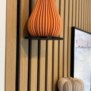 Pode incluir: Vaso decorativo laranja e canelado numa prateleira preta contra uma parede de painéis de madeira. Um objeto menor, branco, em forma de abóbora canelada, está numa prateleira por baixo. Uma imagem emoldurada com a palavra "família" é visível.