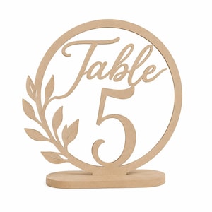 Op de afbeelding: Een lichtbruin houten tafelnummerbord met een cirkelvormig frame, het woord Table en het cijfer 5 in een sierlijk lettertype. Een decoratieve tak met bladeren steekt uit aan de linkerkant van de cirkel en het bord staat op een voetstuk.