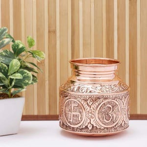 Op de afbeelding: Een koperkleurige, decoratieve pot met ingewikkelde reliëfdesigns. De pot heeft bloempatronen en religieuze symbolen, waaronder een swastika en het Om-symbool. De pot is cilindrisch met een iets bredere basis en een gerolde rand.