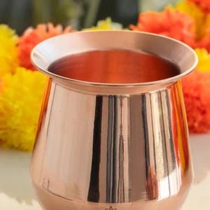 Op de afbeelding: Een gepolijst koperkleurig vat met een brede rand en een afgeronde body. Het vat reflecteert de omgeving. De achtergrond bevat gele en oranje bloemen, wat een levendig contrast met het koper geeft. Het vat wordt waarschijnlijk gebruikt voor decoratieve of ceremoniële doeleinden.