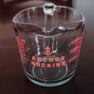 May include: Clear glass measuring cup with a handle and red measurement markings. The cup features the "Anchor Hocking" logo and measurements in cups and liters. A classic kitchen tool for baking and cooking.