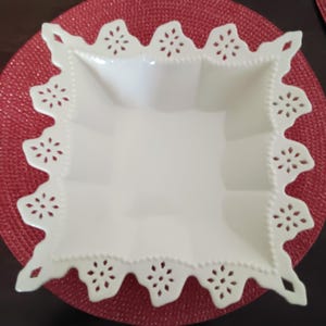 May include: A square, white ceramic serving dish with a decorative, scalloped edge featuring floral cutouts. The dish sits on a red, textured placemat, creating a contrast in color and texture. The dish is empty.