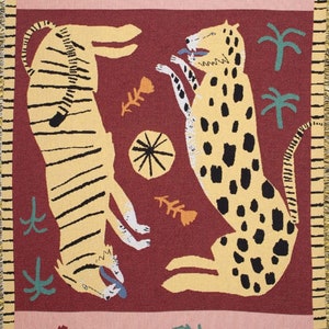 May include: A woven tapestry featuring two stylized animals, a tiger and a leopard, in a playful pose against a burgundy background. The tapestry is framed by a pink border with green floral accents.