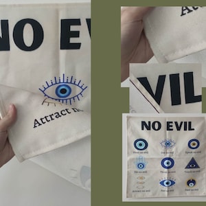 May include: A white fabric wall hanging with the phrase "No Evil" printed in black. The hanging features a blue evil eye design with the text "Attract no evil" printed below. The hanging is also decorated with a series of blue evil eyes with the text "Hear no evil", "See no evil", "Speak no evil", "Do no evil", "Think no evil", "Touch no evil", "Attract no evil", "Feel no evil", and "Feed no evil" printed below each eye.