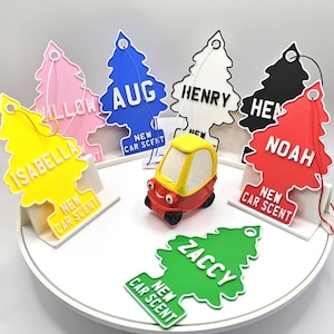May include: A selection of car air fresheners in the shape of pine trees, each bearing a name and the text "New Car Scent." A small red and yellow toy car is positioned in the middle. The air fresheners are in a variety of colours, including pink, blue, white, black, red, yellow, and green.