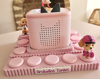 May include: A light pink square audio player with two dark grey cat-ear accents on top, featuring a Moana figurine, a small cartoon character, and a pink puppy figurine. The base is a pink tiered stand with circular slots and a label reading Arabellas Tonies.