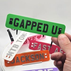 May include: A collection of colorful, rectangular nameplates with various names and phrases. The nameplates are in shades of green, orange, pink, white, and purple, with black or silver lettering. One plate reads "GAPPED UP".