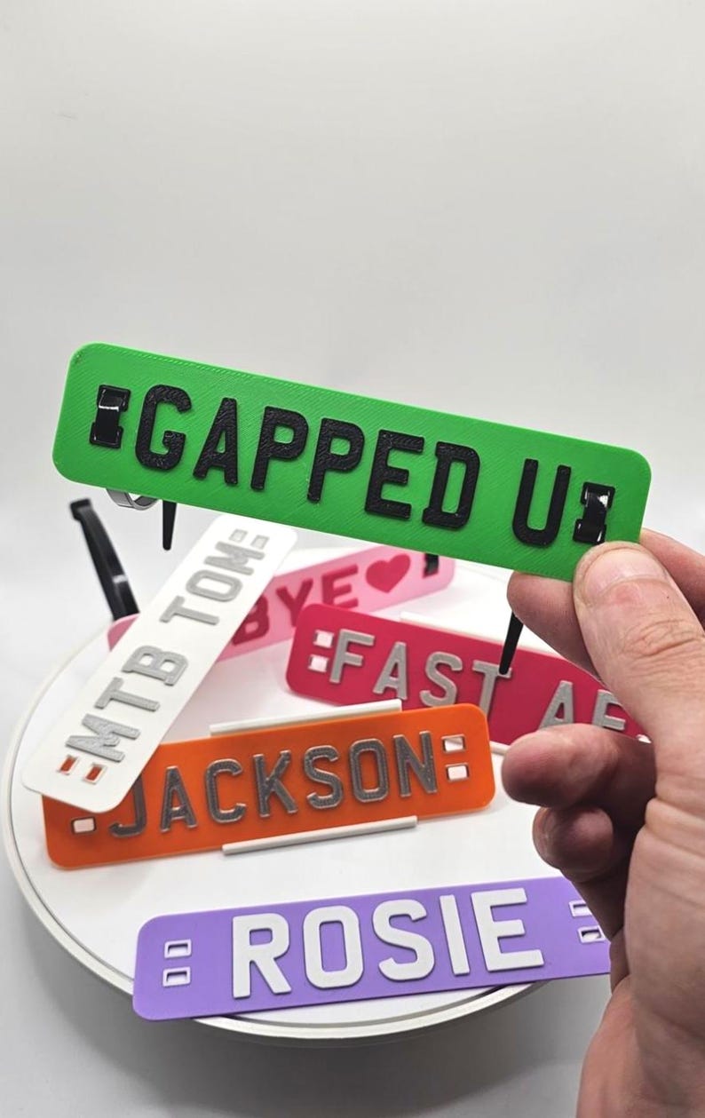 May include: A collection of colorful, rectangular nameplates with various names and phrases. The nameplates are in shades of green, orange, pink, white, and purple, with black or silver lettering. One plate reads "GAPPED UP".