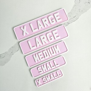May include: A collection of pink rectangular signs with white raised lettering indicating sizes: X LARGE, LARGE, MEDIUM, SMALL, and X SMALL. The signs are arranged diagonally on a light-colored surface.