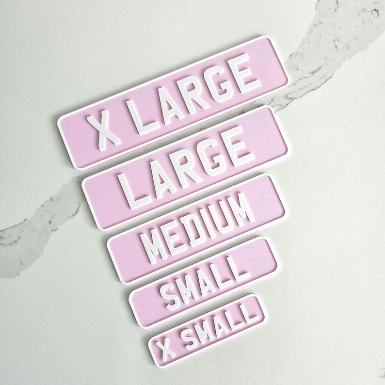 May include: A collection of pink rectangular signs with white raised lettering indicating sizes: X LARGE, LARGE, MEDIUM, SMALL, and X SMALL. The signs are arranged diagonally on a light-colored surface.