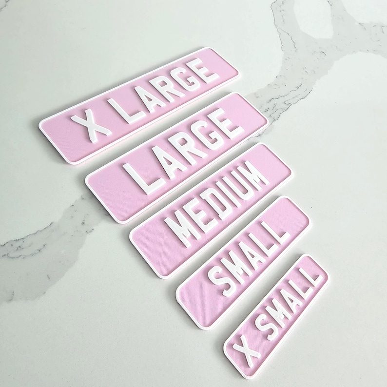 May include: Five pink rectangular signs with white lettering indicating sizes: X LARGE, LARGE, MEDIUM, SMALL, and X SMALL. The signs are arranged diagonally on a light-colored surface.