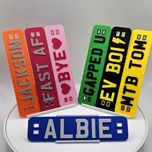 May include: A collection of colorful, rectangular nameplates with various text. The nameplates are orange, red, pink, green, black, and yellow. Text includes "JACKSON", "FAST AF", "BYE", "GAPPED UP", "EV BOIS", and "MTB TOM". A blue nameplate reads "ALBIE".