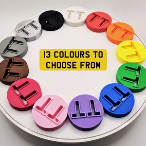 May include: A collection of 13 colorful, circular objects, each featuring a raised "TT" design. The objects are arranged in a circle on a white surface. A yellow rectangle in the center reads "13 COLOURS TO CHOOSE FROM".