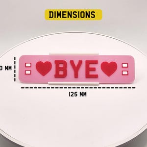 May include: A pink rectangular decorative item with the word "BYE" in red letters, flanked by red hearts. The item is 125 mm long and 30 mm high. Two small white rectangles are on each side of the hearts.