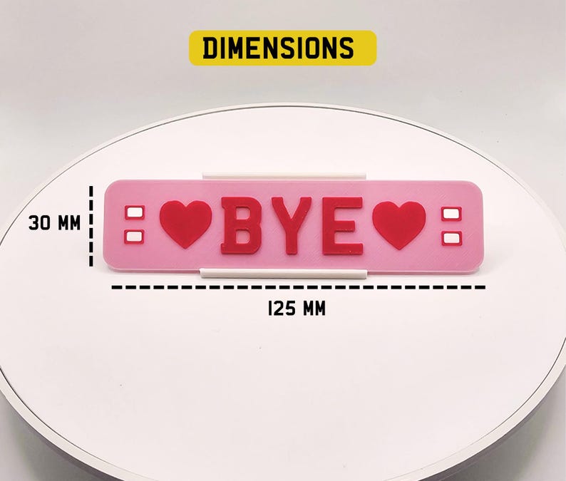 May include: A pink rectangular decorative item with the word "BYE" in red letters, flanked by red hearts. The item is 125 mm long and 30 mm high. Two small white rectangles are on each side of the hearts.