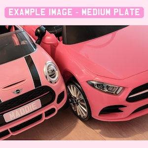 May include: Two pink toy cars are parked next to each other. One is a Mini Cooper with black racing stripes and a license plate that reads "MADDIE". The other is a Mercedes-Benz. The image has the text "EXAMPLE IMAGE - MEDIUM PLATE" at the top.