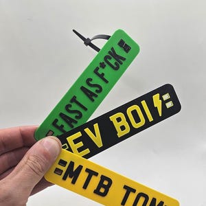 May include: Three rectangular tags with black text. The top tag is green and reads "FAST AS F*CK". The middle tag is black and reads "EV BOI" with a lightning bolt. The bottom tag is yellow and reads "MTB TOM".