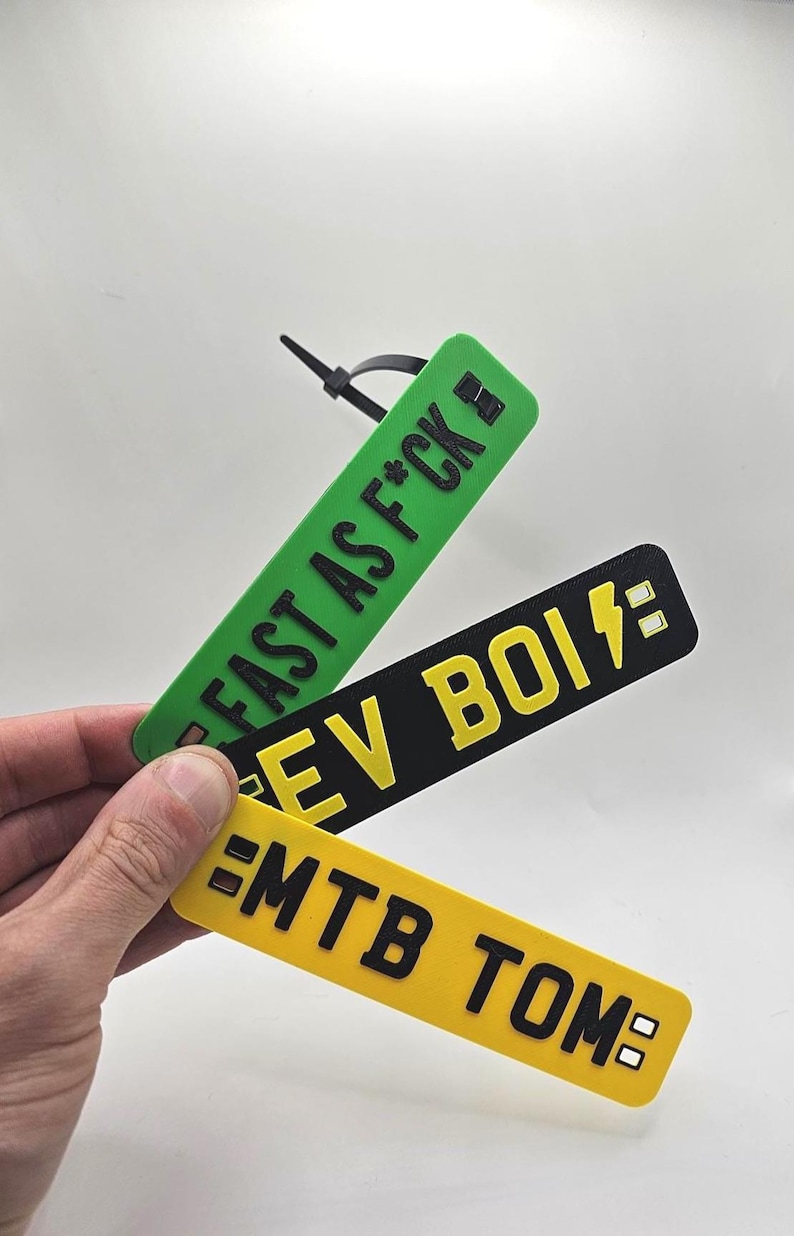 May include: Three rectangular tags with black text. The top tag is green and reads "FAST AS F*CK". The middle tag is black and reads "EV BOI" with a lightning bolt. The bottom tag is yellow and reads "MTB TOM".
